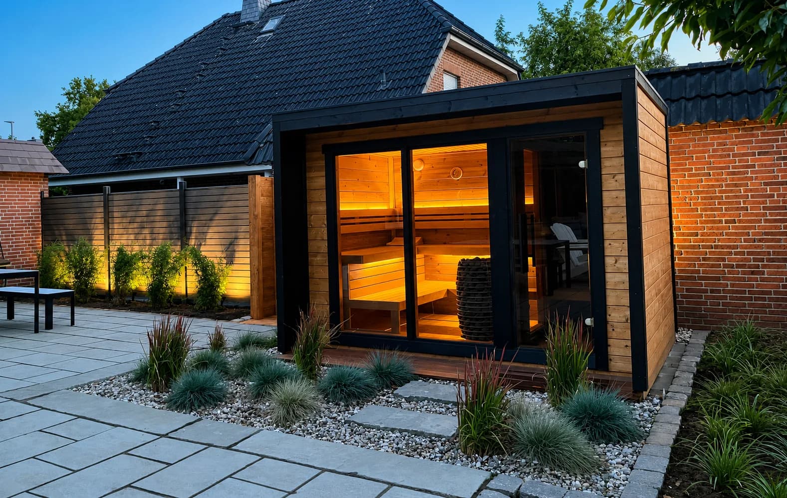 Moderne Outdoor-Gartensauna mit Glasfront und warmer Innenbeleuchtung, eingebettet in eine Gartengestaltung mit Terrassenplatten, Kiesbeeten und Ziergräsern, angelegt von Paul Hermann in Elmshorn